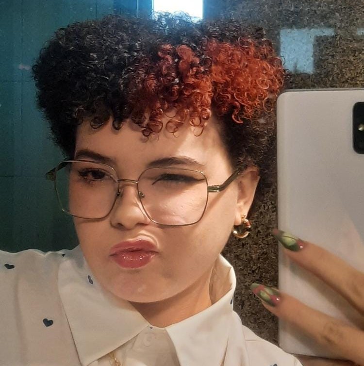 foto da beatriz, uma mulher branca com cabelos crespos pretos com mecha vermelha, usando oculos e uma camisa com gola branca